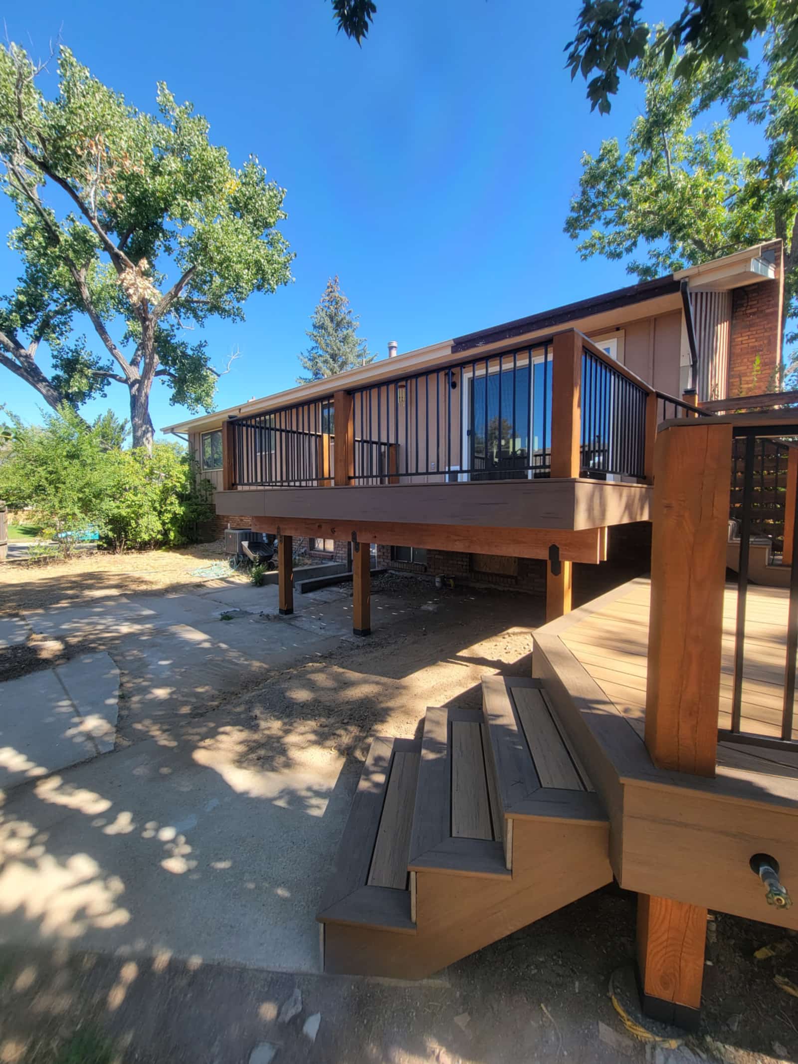 Large cedar deck raised on posts with a wood and black metal railing, mature trees around it