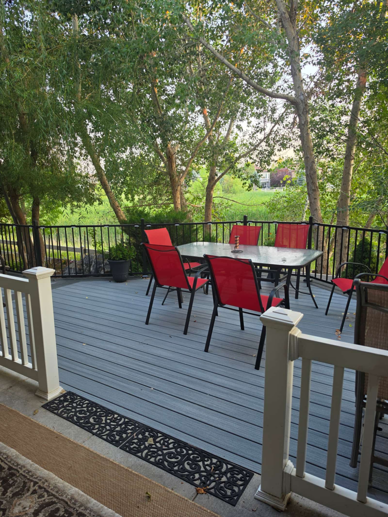 Gray composite deck with black metal railings, furnished with a dining table and red patio chairs under mature trees