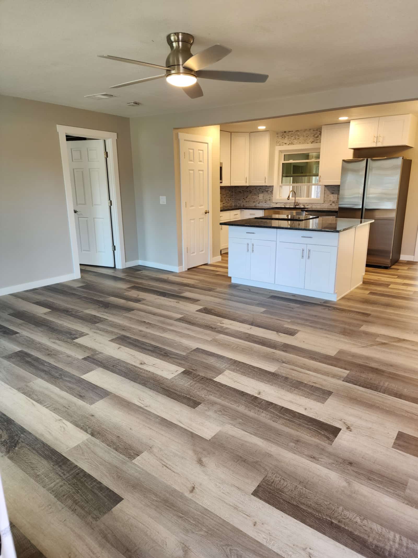 Finished kitchen with white shaker cabinets, dark granite worktops, a mosaic tile backsplash and a central island, opening onto a living space with new plank flooring