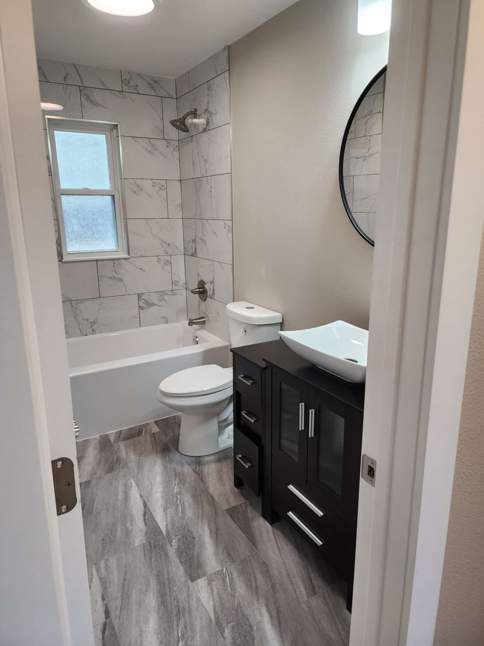Finished bathroom with a marble-look tiled bath surround, a dark vanity with a vessel basin and a round mirror